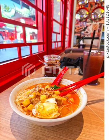 Spicy noodle soup with egg and chicken in vibrant broth. Served with red chopsticks and iced drinks in a cozy cafe setting. Spicy noodle soup with egg and chicken in vibrant broth. Served with red chopsticks and iced drinks in a cozy cafe setting. 131121049