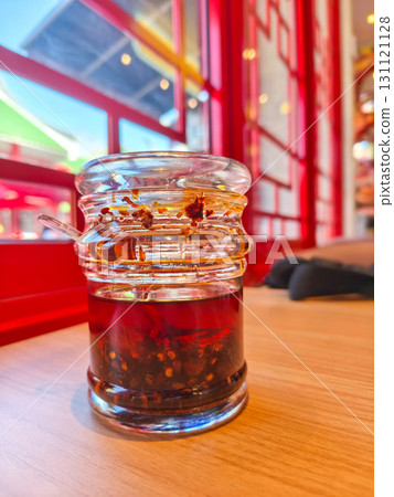 Jar of chili oil with spoon on wooden table in vibrant Asian restaurant. Visible chili flakes and seeds highlight spicy, flavorful condiment. 131121128