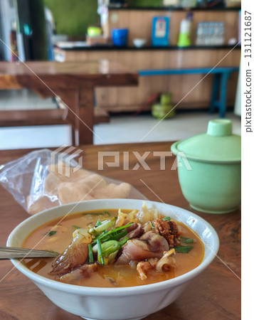 Spicy beef tendon soup with herbs and green onions in rich broth. Served in a white bowl with spoon, accompanied by crackers and a green dish on wooden table. Spicy beef tendon soup with herbs and green onions in rich broth. Served in a white bowl with spoon, accompanied by crackers and a green dish on wooden table. 131121687