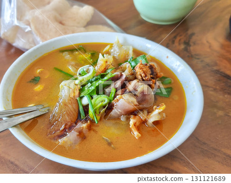 Spicy beef tendon soup with herbs and green onions in rich broth. Served in a white bowl with spoon, accompanied by crackers and a green dish on wooden table. 131121689