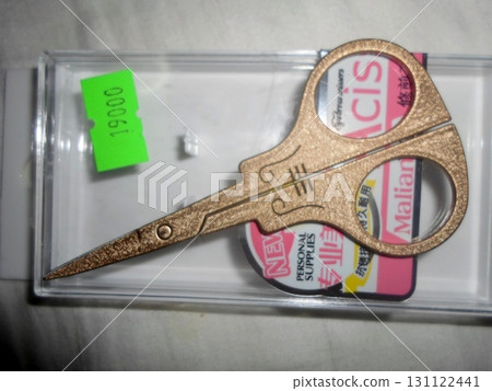 Shiny Gold Scissors in a Clear Plastic Case with a Green Label on a White Surface 131122441