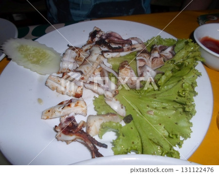 A Plate of Grilled Squid with Lettuce and Cucumber Slices on a Vibrant Yellow Tablecloth 131122476