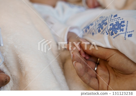 Tender Moment: Parent's Hand Holding Newborn Foot, Soft White Blanket, Blue Embroidered Onesie, Close-Up Shot, Warm Healthcare Scene. Tender Moment: Parent's Hand Holding Newborn Foot, Soft White Blanket, Blue Embroidered Onesie, Close-Up Shot, Warm Healthcare Scene. 131123238