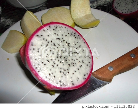 A Sliced Dragon Fruit with White Flesh and Black Seeds on a Plate with a Knife and Apple Slices Nearby 131123828
