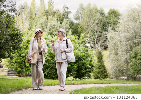 Full length portrait of two senior women walking outdoors in stylish light beige and cream casual outfits with hats, linen trousers, jackets, bag and fashionable accessories in natural park background 131124365