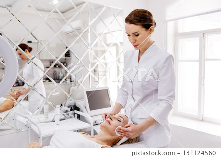Beautician performing facial skincare treatment in modern beauty clinic. Professional cosmetologist cleaning mature woman face with cotton pads Beautician performing facial skincare treatment in modern beauty clinic. Professional cosmetologist cleaning mature woman face with cotton pads 131124560