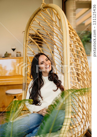 Smiling woman sitting in hanging chair, enjoying a peaceful moment at home Smiling woman sitting in hanging chair, enjoying a peaceful moment at home 131125436