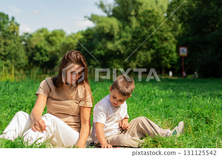 Mother helping her small son reading book about cars and transformers spending time on nature sitting on grass on summer day. 131125542