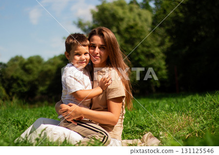 Candid portrait of happy mother and son sitting on green grass at public park on sunny summer day. Holidays with kids. Parenthood and motherhood topics. Candid portrait of happy mother and son sitting on green grass at public park on sunny summer day. Holidays with kids. Parenthood and motherhood topics. 131125546