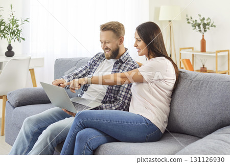 Happy husband and wife at home resting on sofa holdinng laptop to choose goods online, young family  131126930