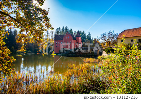 Panoramic view of Cervena Lhota Castle in the Czech Republic, surrounded by a lake reflecting its image. Autumn trees in vibrant colors and sunny weather create a picturesque, serene scene. 131127216