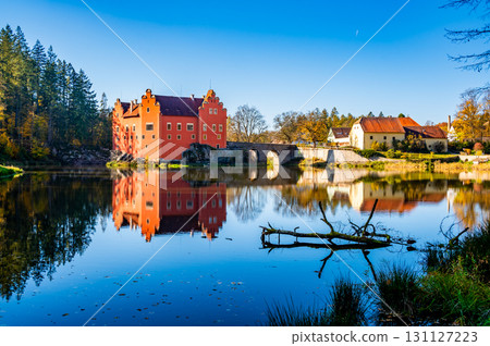 Panoramic view of Cervena Lhota Castle in the Czech Republic, surrounded by a lake reflecting its image. Autumn trees in vibrant colors and sunny weather create a picturesque, serene scene. 131127223