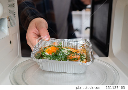 A person's hand places an aluminum takeaway container with food into a microwave, a common kitchen mistake and safety issue. A person's hand places an aluminum takeaway container with food into a microwave, a common kitchen mistake and safety issue. 131127463