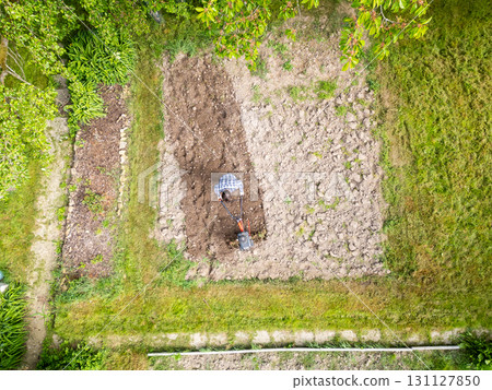 Young farmer man is cultivating the field with machine. Aerial drone top down view of small field in old fashioned garden. Concept of home agriculture. Young farmer man is cultivating the field with machine. Aerial drone top down view of small field in old fashioned garden. Concept of home agriculture. 131127850