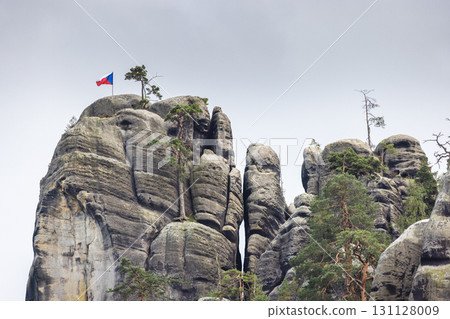 Adrspach-Teplice Rocks, sandstone formations in Hradec Kralove Region in the Czech Republic, Europe. Jagged rock formations rise against a cloudy sky, topped with trees and a fluttering national flag. 131128009