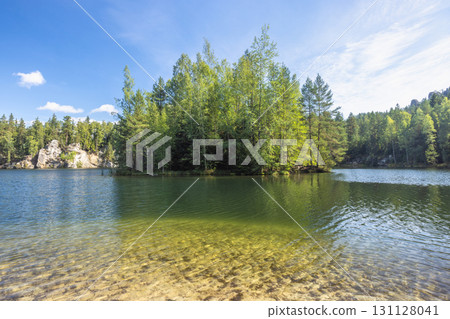The Piskovna lake in Adrspach-Teplice Rocks area, in Hradec Kralove Region in the Czech Republic, Europe. Island in a tranquil lake, surrounded by lush green trees and a clear blue sky above water. The Piskovna lake in Adrspach-Teplice Rocks area, in Hradec Kralove Region in the Czech Republic, Europe. Island in a tranquil lake, surrounded by lush green trees and a clear blue sky above water. 131128041