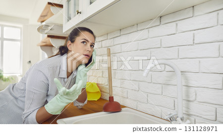 Young girl talking by the phone advising how to unclog a sink drain in the kitchen at home. Young girl talking by the phone advising how to unclog a sink drain in the kitchen at home. 131128971