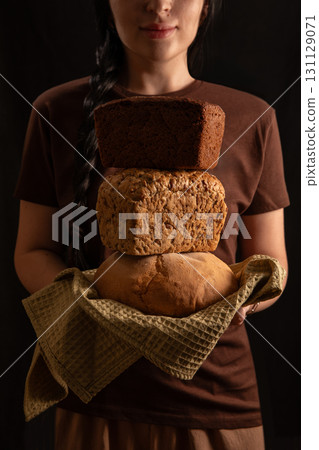 brunette girl stands with a braid proudly holding three types of freshly baked bread wrapped in a kitchen towel. She wears a simple brown T shirt creating a cozy atmosphere. 131129071
