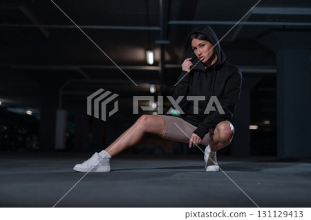 In a dimly lit underground parking lot a girl in a black hoodie squats with one leg extended. She exudes confidence and style while showcasing casual street fashion. 131129413
