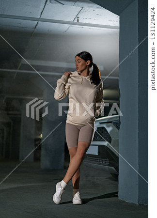 girl wearing a beige hoodie and light shorts strolls through an underground parking lot surrounded by a calm atmosphere. Soft lighting highlights her presence as she walks confidently. 131129424
