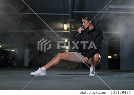 girl wearing a black hoodie crouches in a dimly lit underground parking lot. The atmosphere is enhanced by the surrounding fog and soft lights. She appears contemplative and stylish. girl wearing a black hoodie crouches in a dimly lit underground parking lot. The atmosphere is enhanced by the surrounding fog and soft lights. She appears contemplative and stylish. 131129425