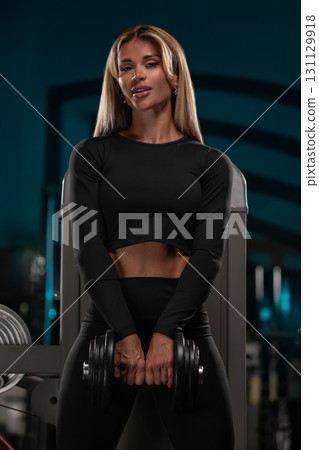 blonde woman works out at a gym in the evening. She holds dumbbells and focuses on her strength training routine. Her outfit is comfortable and suited for exercising. 131129918
