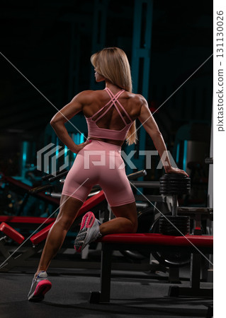 blonde woman is exercising at the gym positioned near weights. She wears a pink outfit and appears strong as she focuses on her workout. The gym has a modern sleek design. 131130026