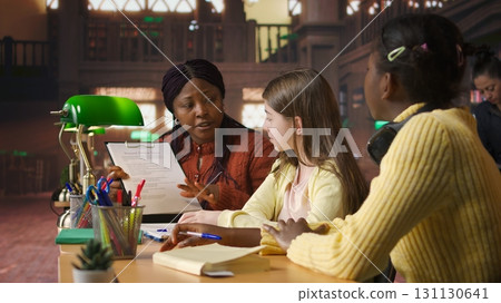 African american professor mentoring diverse schoolgirls in the library, learning together in a library filled with curriculum based materials. Woman tutoring pupils on literature. Camera A. African american professor mentoring diverse schoolgirls in the library, learning together in a library filled with curriculum based materials. Woman tutoring pupils on literature. Camera A. 131130641