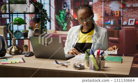 Remote worker at home office desk looking over folder files and using laptop, drafting contracts. Remote employee checking paperwork documents in front of notebook, camera A 131130664