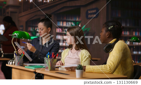 Elementary school pupils working with a professor solving exercises for the class homework, developing learning skills at a study desk. Tutor explains the curriculum materials to girls. Camera B. Elementary school pupils working with a professor solving exercises for the class homework, developing learning skills at a study desk. Tutor explains the curriculum materials to girls. Camera B. 131130669