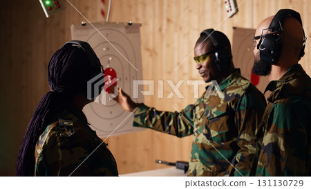 Portrait of joyous army soldiers wearing safety gear in shooting gallery, preparing to fire at targets. Smiling military units in uniform and protection equipment testing accuracy, camera B 131130729
