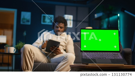 Young black adult uses his tablet next to green screen in his cozy modern apartment, laying on the couch and relaxing with his gadgets at home. Person with neon lights space, chill guy. Camera B. 131130774