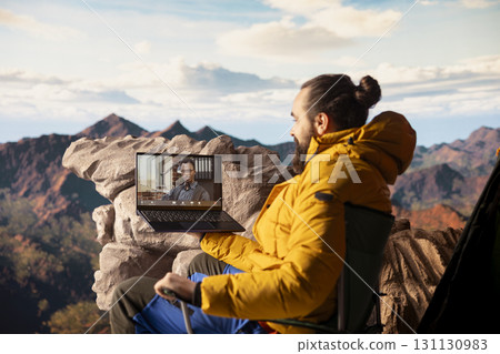 Young CEO entering a video call from the mountain top with a strong signal, always connected and discussing with associates about business matters. Workaholic businessman hiking. 131130983