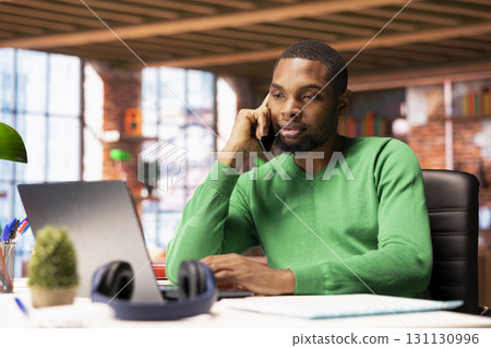 Freelancer in home office having important phone call with business partner using smartphone. African american person sitting at desk discussing company matters during telephone call Freelancer in home office having important phone call with business partner using smartphone. African american person sitting at desk discussing company matters during telephone call 131130996