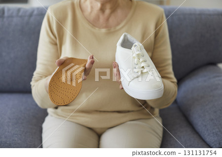 Senior woman sitting on sofa at home with orthopedic insole and sneakers in hands. 131131754