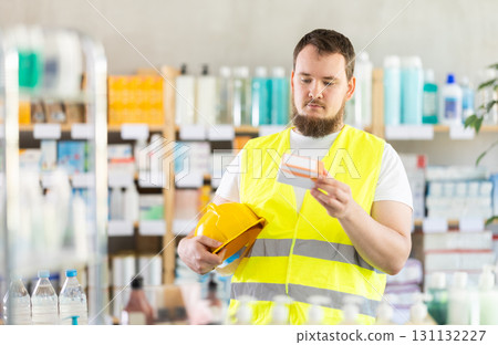 Male builder in yellow vest with helmet in his hands looks for medicine in pharmacy 131132227