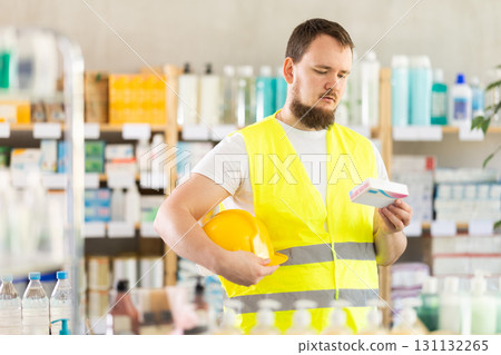 Male builder in yellow vest with helmet in his hands looks for medicine in pharmacy Male builder in yellow vest with helmet in his hands looks for medicine in pharmacy 131132265
