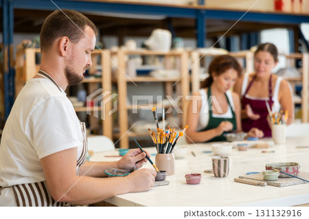 Male potter learns how to make designs on clay plate using brush and paint in pottery workshop 131132916