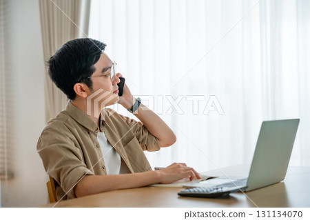 Happy young Asian man working on laptop in living room Happy young Asian man working on laptop in living room 131134070
