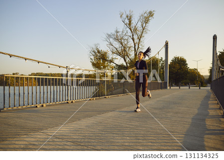 Young slim sporty woman running across a bridge in a city park. Girl in sportswear jogging outdoor 131134325