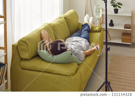 Young man receiving IV drip infusion and vitamin therapy lying on the couch at home. 131134328