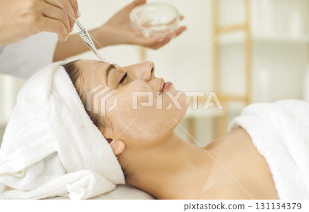 Female cosmetologist applying moisturizing mask with brush to a young woman in spa salon. 131134379