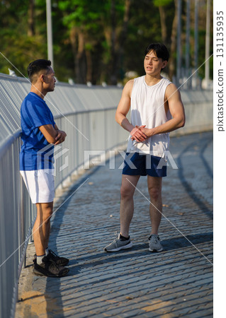 Two asian men or male friends talking together while standing at sports stadium fence or railing. 131135935