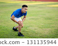 Blue shirt asian man is crouching and holding his knee while standing on grass field. Pain or injury 131135944