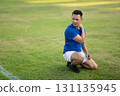 An asian man in blue t shirt is touching his shoulder and looking away while kneeling on grass field 131135945