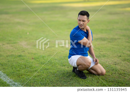 An asian man in blue t shirt is touching his shoulder and looking away while kneeling on grass field 131135945