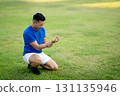 A man in blue shirt holding and looking at his wrist while sitting on grass field. Pain or injury. 131135946