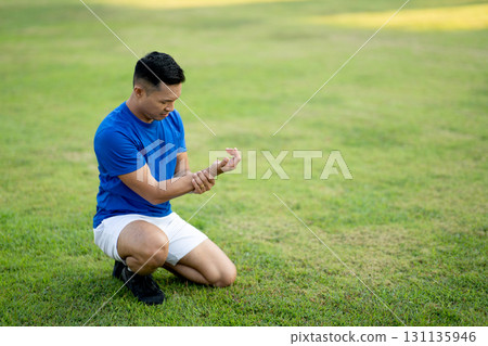 A man in blue shirt holding and looking at his wrist while sitting on grass field. Pain or injury. 131135946