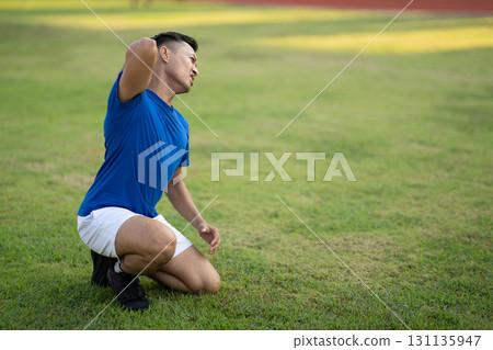 An asian man in blue t shirt touching his back while kneeling on the grass field. Pain or injury. 131135947