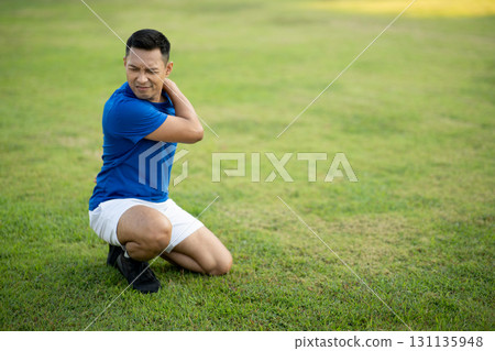An asian man in blue t shirt is touching his shoulder while kneeling on grass field. Pain or injury. 131135948